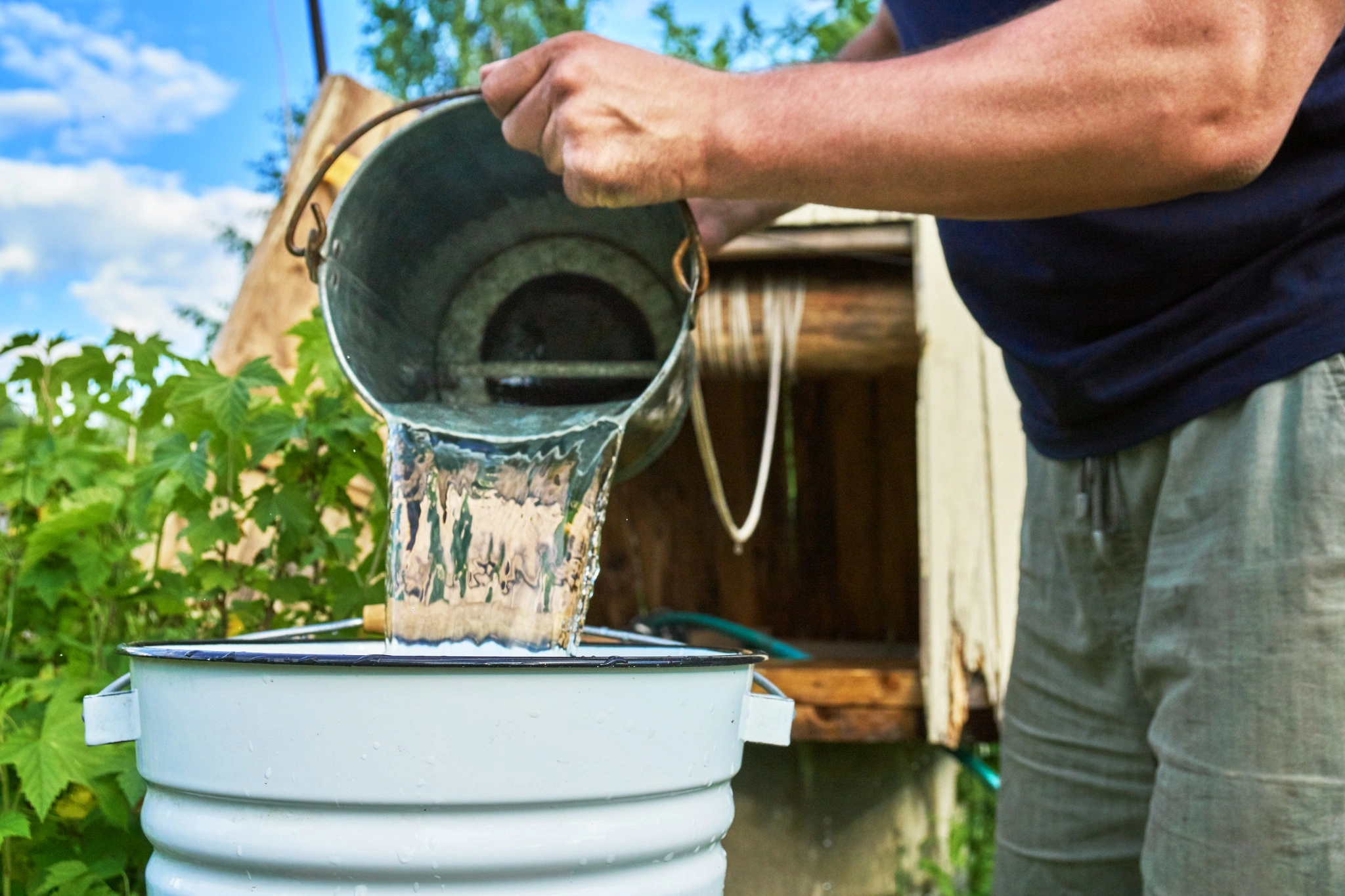 Person pouring well water from a metal bucket into a basin outdoors, illustrating summer well water safety and bacteria risks in New Hampshire