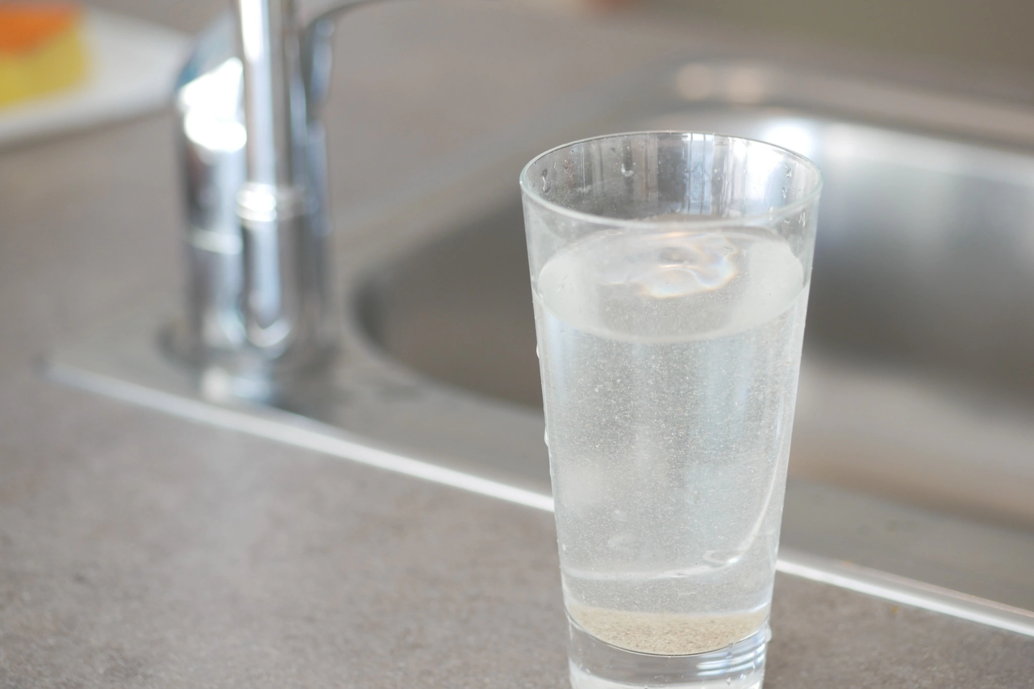 Glass of tap water with visible sediment at the bottom near a kitchen sink illustrating potential issues from ignoring water testing results
